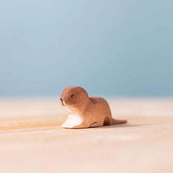 The Bumbu Handmade Wooden Baby Otter, an eco-friendly toy, sits charmingly on a light wooden surface against a soft blue background.