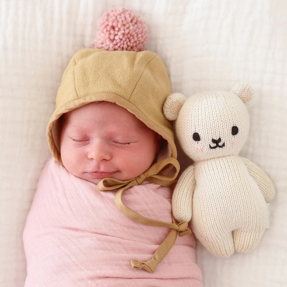 A sleeping baby wrapped in a pink blanket is lying on a white quilted surface. The baby, wearing a beige bonnet with a pink pom-pom, rests beside a Cuddle + Kind Baby Polar Bear.