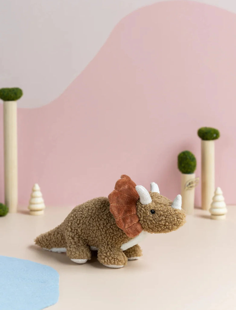 A cute Baby Triceratops Dinosaur Stuffed Animal is placed on a flat surface with a pastel pink and blue backdrop, decorated with small artificial trees and foliage in the background. This dinosaur toy has a brown, textured body with distinctive white horns and a rust-colored frill, suitable for infants.