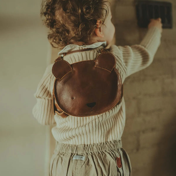 A toddler from behind wearing a cream sweater and gray pants, holding onto a railing, with a cute brown bear-shaped Donsje Mini Leather Backpack - Bear.