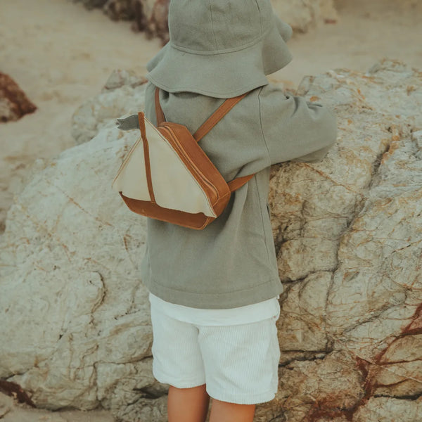 A young child in a hoodie and shorts, seen from behind, stands by rugged rocks at the beach, wearing a small Donsje Mini Leather Backpack - Boat.