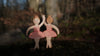 Two Handmade Holzwald Ballerina toys in pink tutus stand on a mossy forest floor, one raising an arm and holding the other's hand, inviting imaginative play among blurred trees and brown foliage.