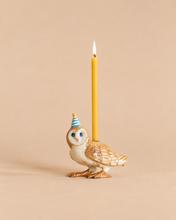 A handcrafted Barn Owl Cake Topper, featuring a ceramic owl wearing a blue party hat and holding a lit yellow candle on its back, is showcased against a plain beige background.