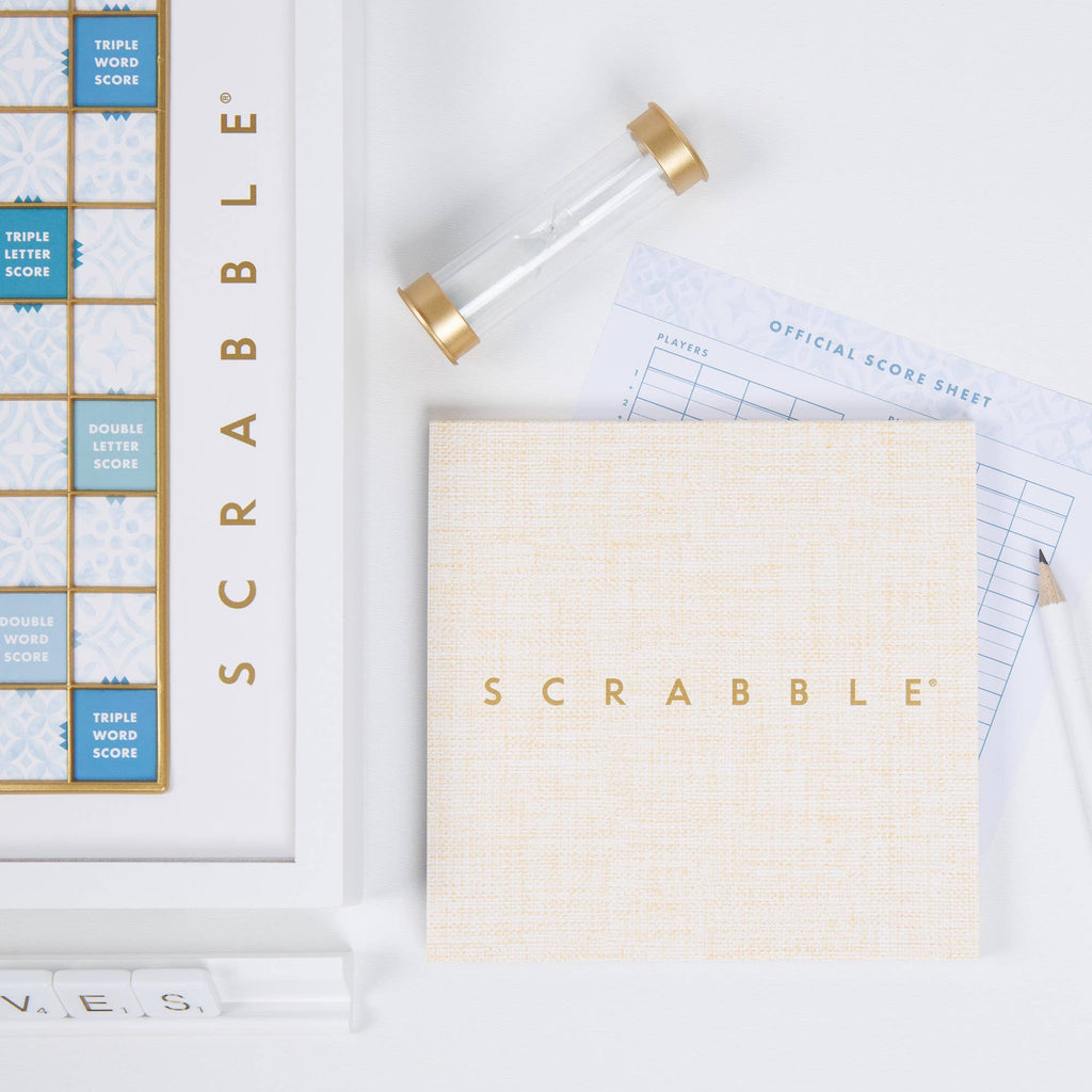 A Scrabble Bianco Edition board game set featuring a pale blue and white wood-framed board, a sand timer, a score sheet, and a square box lid with a textured tan cover.