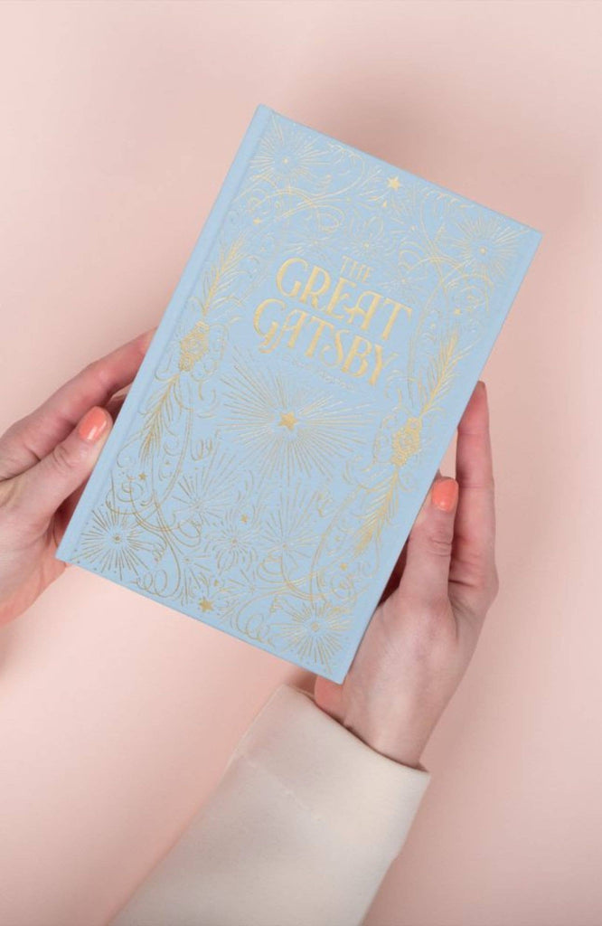 A person with peach-colored nails holds The Great Gatsby | Fitzgerald | Luxe Edition | Hardcover, a light blue book with gold embossed designs, against a pale pink background.