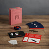 The Board Game: Scattergories Vintage Bookshelf Edition is displayed on a wooden table, featuring a pink book-shaped box, game pads, cards, timer, pens, large white die, and instruction sheets inspired by the 1988 original.