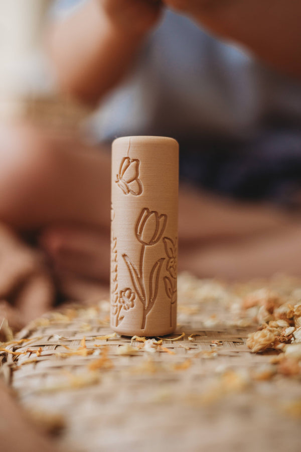 The Flower Roller - Eco Pattern Playdough Roller, featuring engraved tulip and butterfly designs on a wooden cylinder, stands upright on a woven mat surrounded by small dried petals—a perfect eco-friendly tool for sensory play.