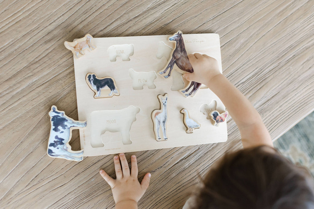 A child places animal-shaped pieces from the Farm Animals Wooden Puzzle: A Tabitha Paige Puzzle onto a wooden board, matching the images and names of animals such as cow, llama, and dog. The colorful puzzle serves as an educational toy for toddlers, with the child’s hand and puzzle pieces visible on a wooden table.