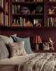 A cozy reading nook features maroon bookshelves, a patterned bench, and cheerful decor. Highlighted is the Coral & Tusk Tree of Fun Pillow, adding playful charm near a lamp, animal figurines, and children's wooden toys.