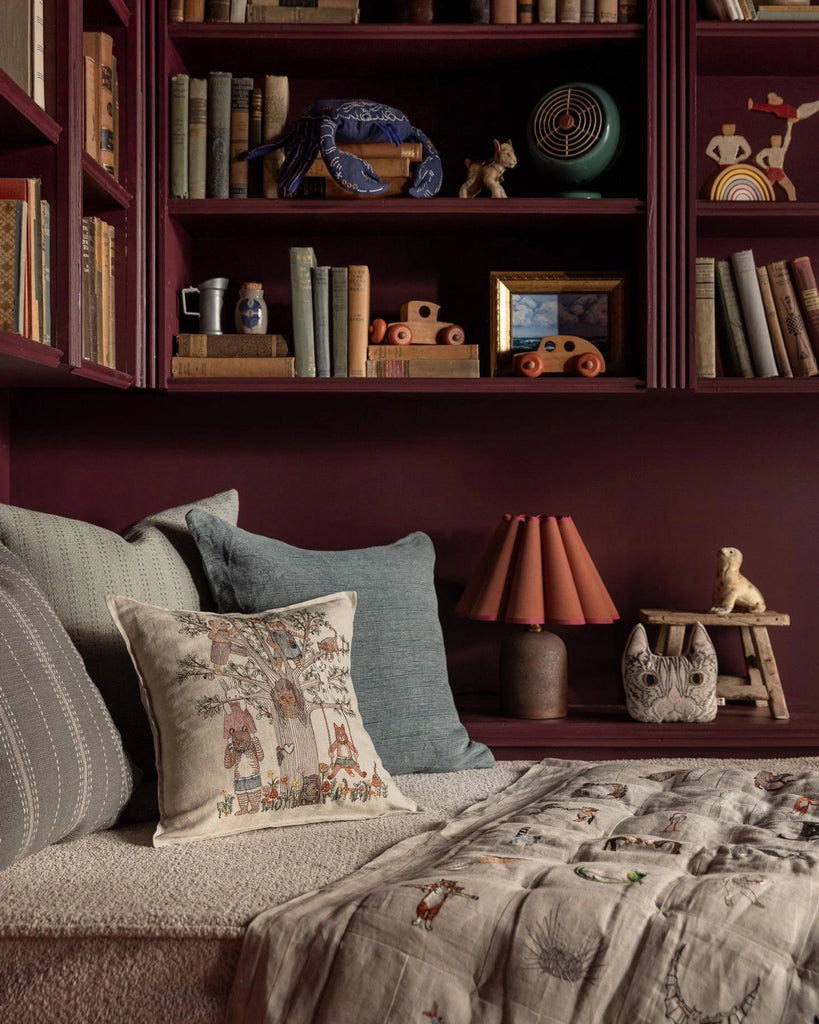 A cozy reading nook features maroon bookshelves, a patterned bench, and cheerful decor. Highlighted is the Coral & Tusk Tree of Fun Pillow, adding playful charm near a lamp, animal figurines, and children's wooden toys.