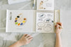 A child uses the Wishy Washy (Water Reveal Edition) on a marble surface, revealing animal-themed activity pages with a paint palette and sound-making animals—perfect as a reusable activity book for toddlers.