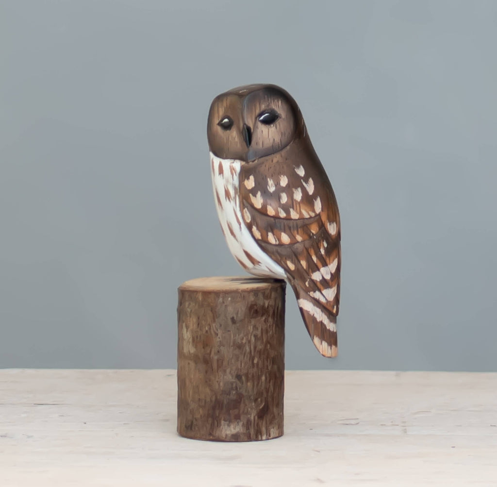 The Hand Carved Ornament - Mini Barred Owl - 5”H is crafted from sustainable softwood, painted by hand, and displayed on a short round log against a plain light gray background.