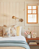 A cozy bedroom features white wood panel walls, a brass wall lamp, a small wooden nightstand with pink tulips, stacked books, and the Coral & Tusk Easter Bunny Pocket Pillow on the bed with other floral cushions under the window.