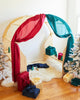 The wooden playhouse, dressed in a giant Sarah's Silk Evergreen playsilk with red accents, is surrounded by a collection of vibrantly wrapped gifts and rests on a soft white rug. On either side of the scene, two small frosted Christmas trees contribute to the festive ambiance with their sophisticated winter hues.