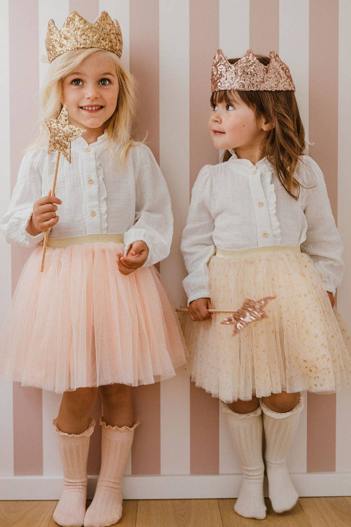 Two young girls, one smiling at the camera and the other looking at her, stand in front of a pink and white striped wall. Both wear gold crowns, white blouses, pastel tulle skirts, hold star wands, and one wears the Cape - Pink sequins.