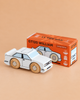 A white No. 21 GT120 William wooden toy car by Le Toy Van is displayed in front of its orange box packaging labeled “GT120 WILLIAM Limited Edition.” Set against a solid tan background, the scene captures the nostalgic appeal of a classic English wooden racing car.