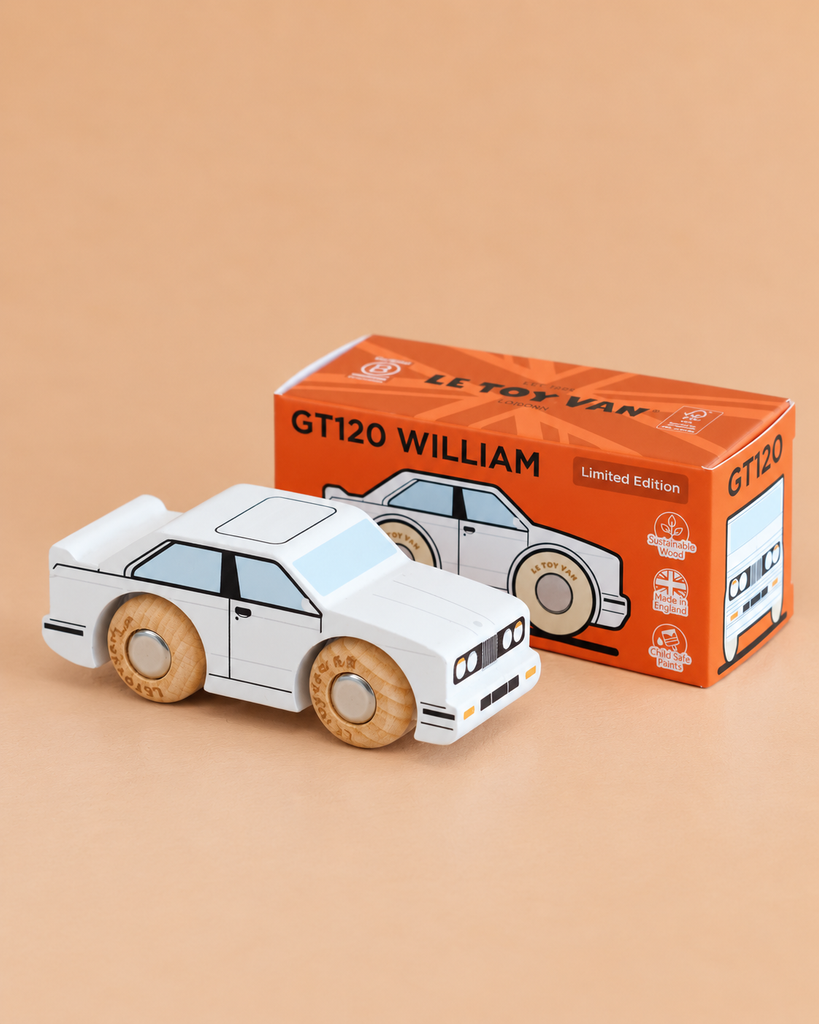 A white No. 21 GT120 William wooden toy car by Le Toy Van is displayed in front of its orange box packaging labeled “GT120 WILLIAM Limited Edition.” Set against a solid tan background, the scene captures the nostalgic appeal of a classic English wooden racing car.