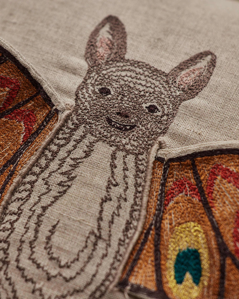Close-up of the Coral & Tusk Bat Wing Surprise Pillow, showcasing detailed embroidery of a smiling brown bat with large ears and vibrant butterfly wings—an enchanting touch for whimsical Halloween decor or costume inspiration.