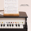 The Wooden Grand Piano Toy features colored note labels on its keys, a music sheet on a wooden stand, and is made from FSC-certified wood. The right side of the image displays “Learn to Play With Colours or Notes.”.