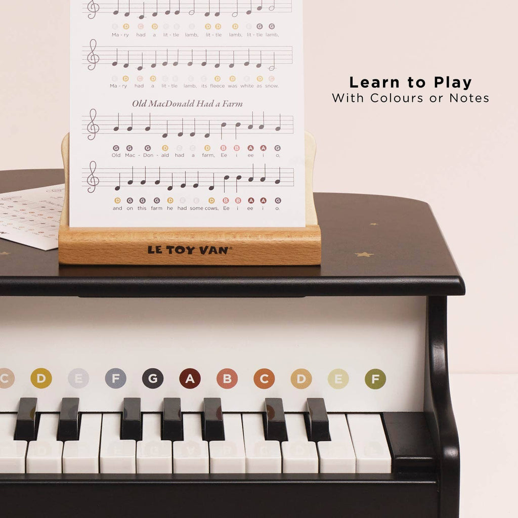 The Wooden Grand Piano Toy features colored note labels on its keys, a music sheet on a wooden stand, and is made from FSC-certified wood. The right side of the image displays “Learn to Play With Colours or Notes.”.