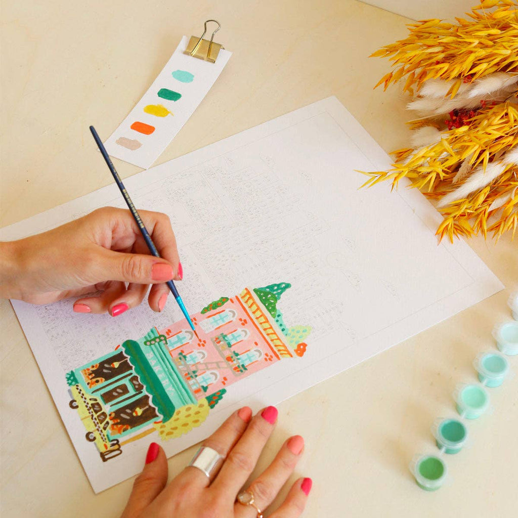 With pink nails, a person uses a brush to complete the Paint By Number Kit - New York. Paint pots, a swatch card, and yellow dried flowers decorate the table.
