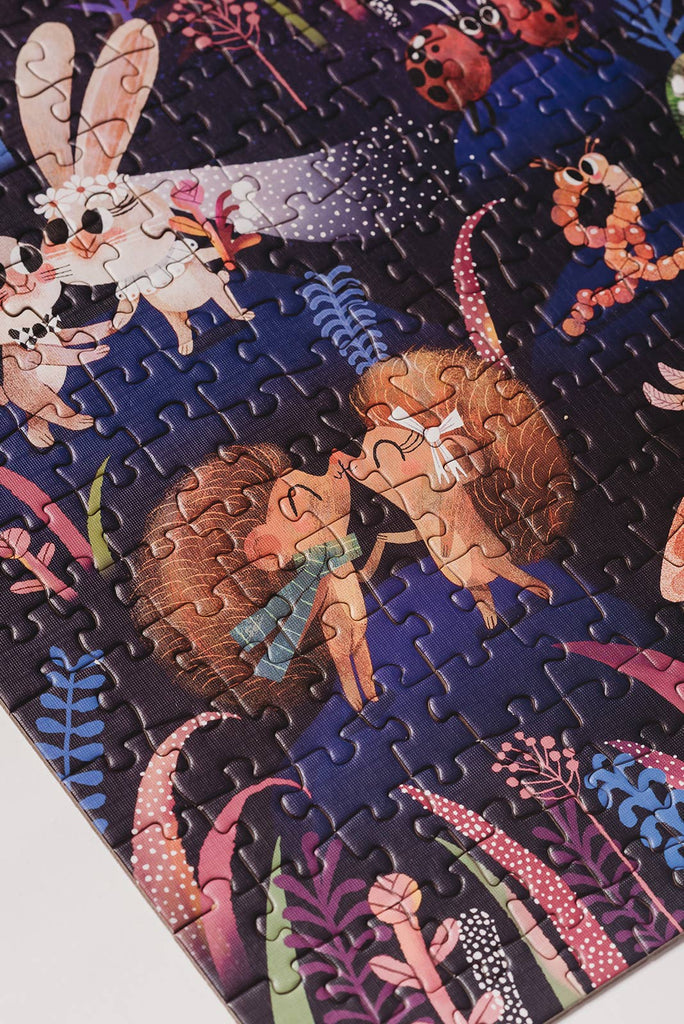 A close-up of the Courting Critters 500 Piece Puzzle reveals illustrated animals—two hedgehogs touching noses, a white rabbit, and a caterpillar—nestled among vibrant plants on a dark background. This charming puzzle is perfect for adults and makes a wonderful Valentine’s Day gift for someone special.