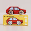A classic red wooden car, No. 8 GT50 Gary, with matching wooden wheels sits atop a yellow box labeled “GT50 GARY.” The box showcases an illustration of the toy along with icons indicating sustainable wood, made in England, and child-safe paint. With its retro cool and vintage charm, this Made in England wooden car is a delightful addition to any collection.