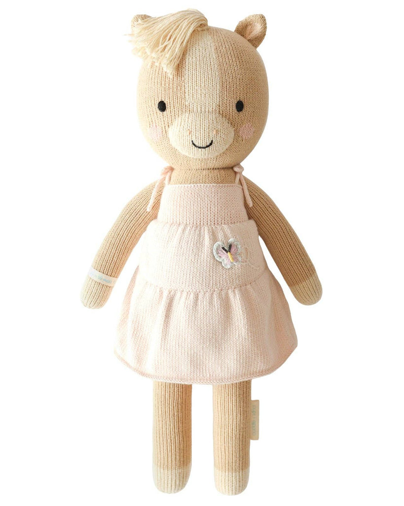 Cuddle + Kind Kelsie the Pony is a hand-knit, fair trade doll made in Peru. It features beige yarn, a cream mane, a pink dress with butterfly detail, embroidered eyes, a smiling mouth, and blushing cheeks.