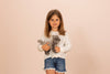 A young girl with long brown hair wearing a white polka-dotted sweater and denim shorts stands against a plain background. She is holding a Teddy Hermann Gray Standing Cat Stuffed Animal. She looks directly at the camera with a neutral expression, showcasing its child-friendly design.