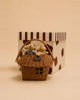 The "Little Forest Animals — The Thompson Family" rattan basket gift set, featuring three plush teddy bears, sits in front of a large cream box adorned with brown spots and stripes, all against a beige background.