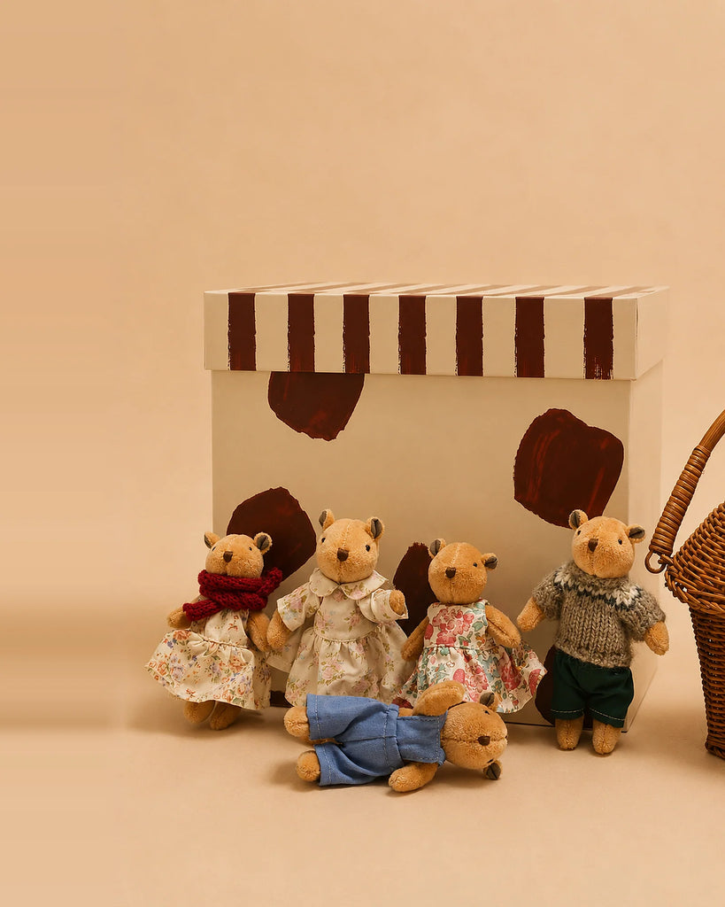 Five handcrafted Little Forest Animals — The Thompson Family dolls, dressed in charming outfits, are posed in front of a cream box with brown stripes and shapes. A rattan basket gift set is placed to the right. The background and floor are light brown.