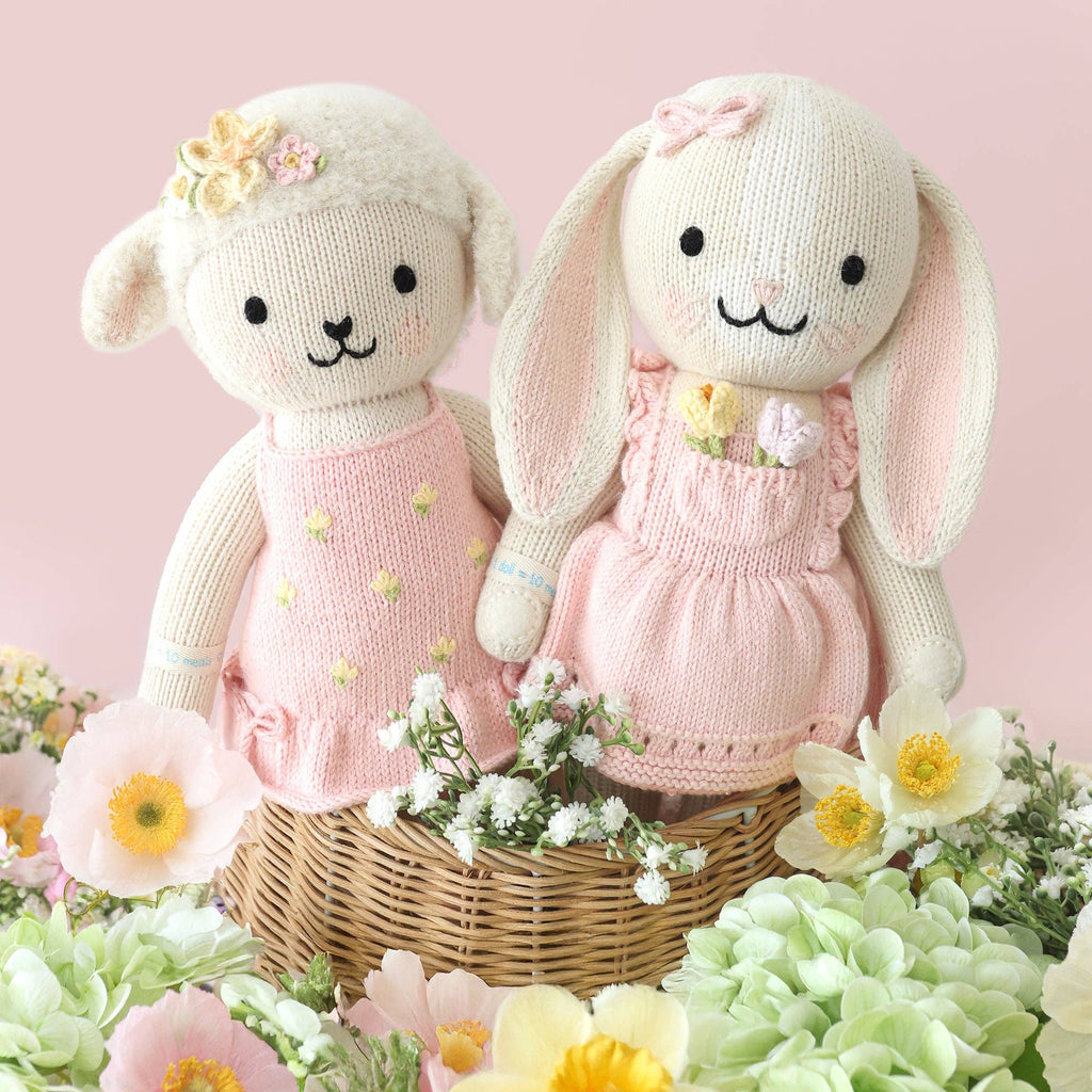 Cuddle + Kind Clover the Bunny (Blossom), a fair trade hand-knit stuffed animal in a pink dress, sits in a wicker basket with pastel flowers and greenery, brightening the soft pink background.
