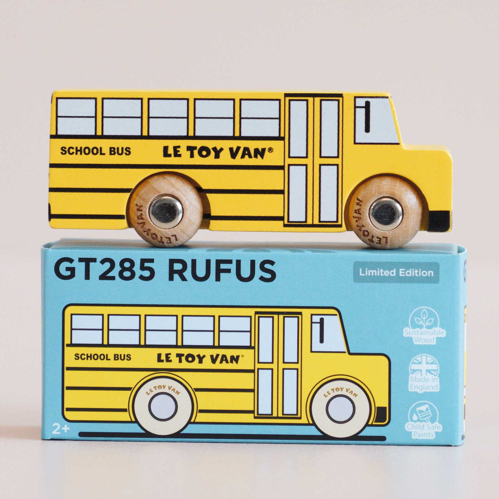 A retro wooden school bus painted in vibrant yellow with “LE TOY VAN” branding sits atop a blue box labeled “No. 47 GT285 Rufus | Made in England.” The box features an illustration matching the toy bus, capturing classic American charm and inviting imaginative journeys. Text on the packaging indicates it is a limited edition, perfect for ages 2 and up.