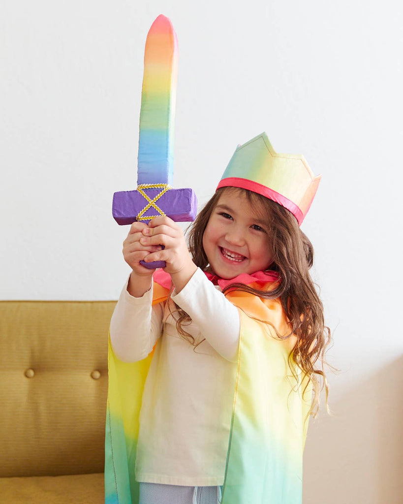 A child beams with joy, gripping a Sarah's Silk Soft Sword in a rainbow color. Dressed in a matching rainbow cape and crown, they stand before a plain white wall, embodying the spirit of imaginative pretend play and active adventure.