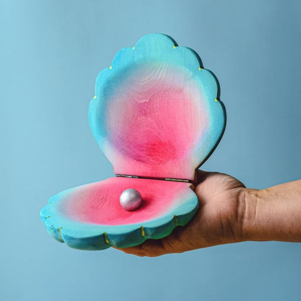 A hand holds the Bumbu Handmade Wooden Big Shell with Pearl, painted in blue, pink, and purple, with a small pearl inside, set against a light blue background.