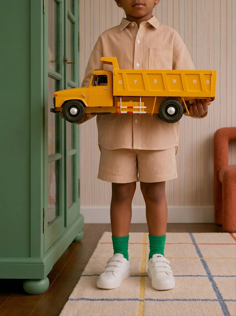 A child wearing a striped beige shirt and shorts with green socks and white shoes stands in a colorful, neatly decorated room, holding the Little Forest Animals — Big Yellow Truck.