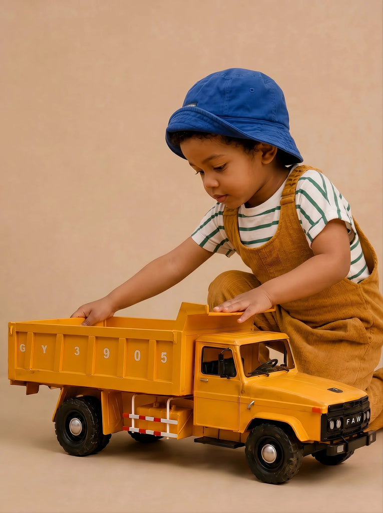 A young child in a blue hat and brown overalls plays with Little Forest Animals — Big Yellow Truck, a handcrafted toy truck inspired by a large yellow FAW dump truck model, against a plain beige background.