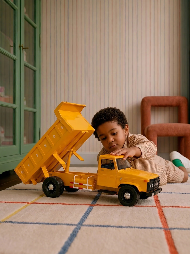 A young child lies on a rug, playing with the Little Forest Animals — Big Yellow Truck in a cozy room with striped wallpaper and colorful furniture.