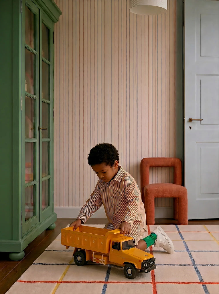 A young child kneels on a patterned rug, playing with the Little Forest Animals — Big Yellow Truck in a cozy room featuring a green cabinet, peach chair, and light blue door.