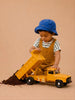A young child in a blue hat and brown overalls plays with the Little Forest Animals — Big Yellow Truck, unloading dirt onto the floor against a plain beige background.