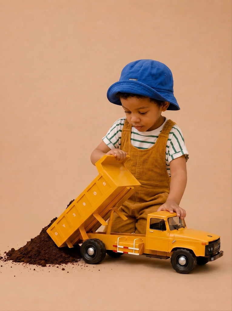 A young child in a blue hat and brown overalls plays with the Little Forest Animals — Big Yellow Truck, unloading dirt onto the floor against a plain beige background.