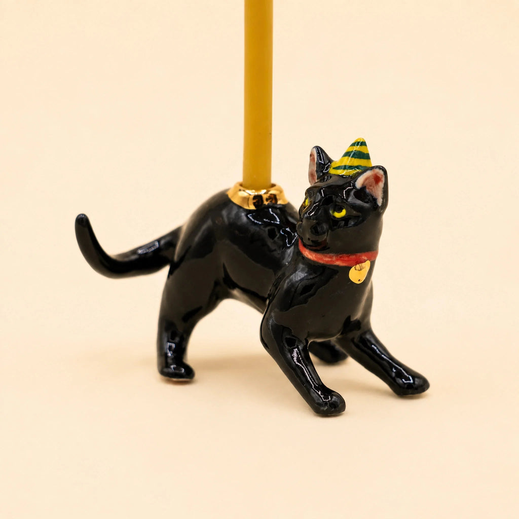 Introducing the Black Cat Cake Topper - Final Sale: This hand-painted porcelain collectible features a black cat crouched with its tail up, wearing a small green and yellow party hat and a red collar adorned with a gold bell. The cat holds a yellow candle on its back, making it a charming cake topper and perfect keepsake birthday gift.