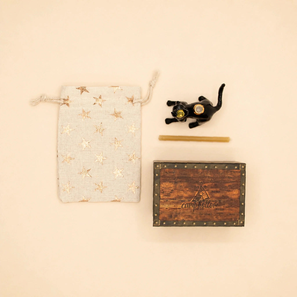 A Black Cat Cake Topper - Final Sale, hand-painted and holding binoculars, is displayed alongside a gold star drawstring pouch, a slender stick, and a rustic wooden box labeled “camptellow,” all set on a beige background.