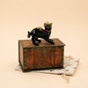 The Black Cat Cake Topper - Final Sale is a small, hand-painted collectible figurine featuring a black cat in a yellow party hat, standing atop a wooden chest with brass accents, all presented on a beige drawstring pouch adorned with a subtle star pattern.
