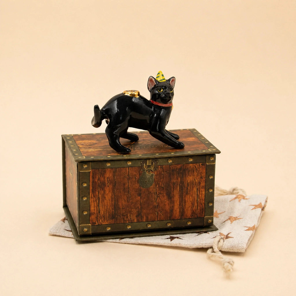 The Black Cat Cake Topper - Final Sale is a small, hand-painted collectible figurine featuring a black cat in a yellow party hat, standing atop a wooden chest with brass accents, all presented on a beige drawstring pouch adorned with a subtle star pattern.