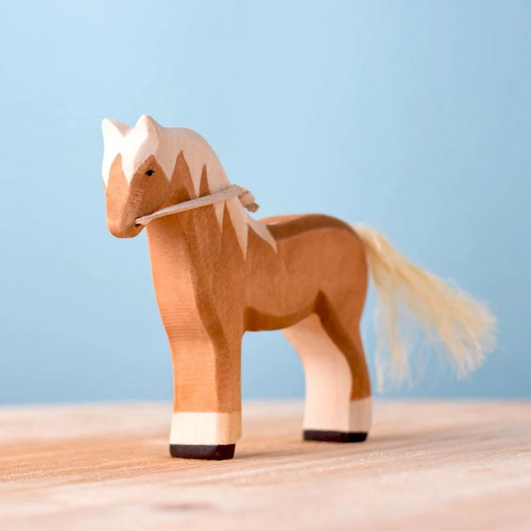 The Bumbu Handmade Wooden Blonde Horse, featuring a light brown body, white mane and tail, and white leg accents, stands on wood against a light blue background—ideal as an heirloom toy for imaginative play.