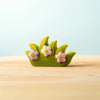 The Bumbu Handmade Wooden Blossom Bush is a small wooden decoration shaped like green grass with three pastel flowers—pink, white, and purple—that evokes natural charm. This Montessori toy rests on a light wooden surface against a pale blue background.