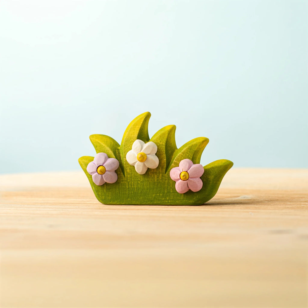 The Bumbu Handmade Wooden Blossom Bush is a small wooden decoration shaped like green grass with three pastel flowers—pink, white, and purple—that evokes natural charm. This Montessori toy rests on a light wooden surface against a pale blue background.