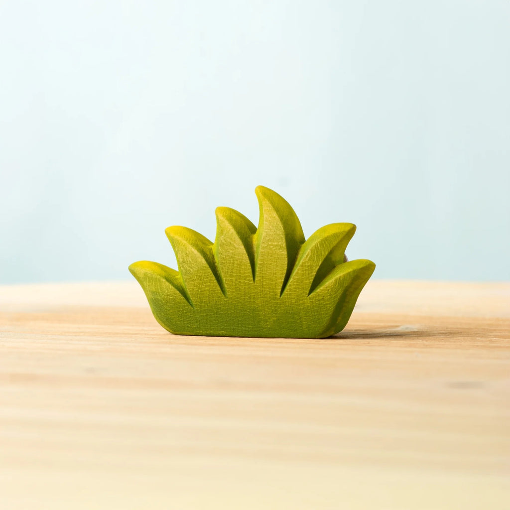 A Bumbu Handmade Wooden Blossom Bush, small and green with a grass-like shape, sits on a light wooden surface against a pale blue background.