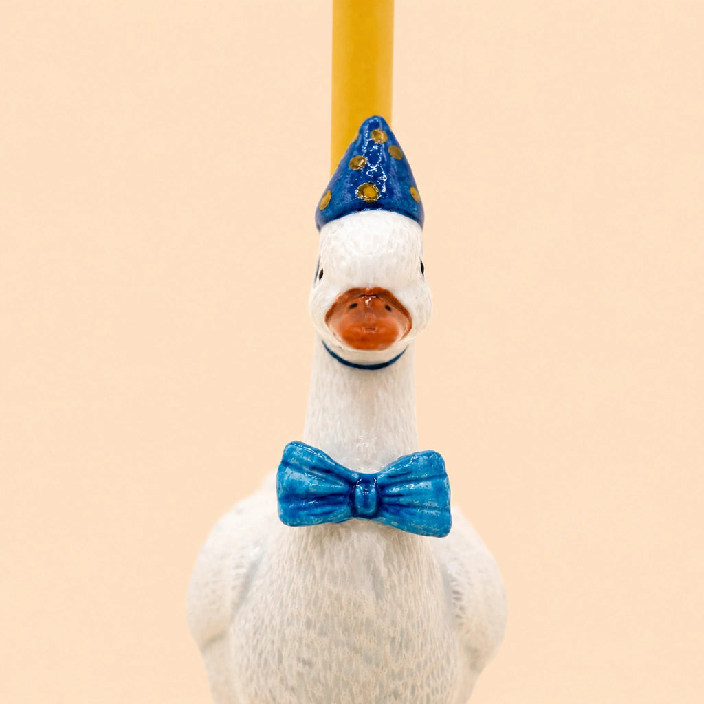 The Blue Goose Cake Topper - Final Sale is a handcrafted birthday decoration featuring a white goose figurine dressed in a blue bow tie and a blue party hat adorned with yellow dots, set against a beige background.